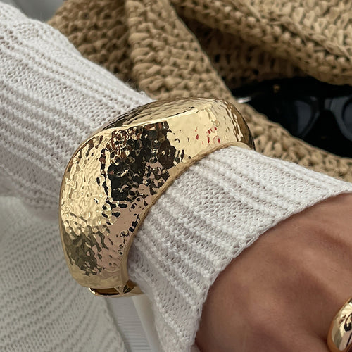 Gold bracelet on a wrist with a textured surface, worn over a white sleeve.