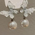 Silver earrings with clear gemstones and pearls held by a hand against a neutral background