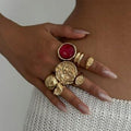 Hand wearing multiple gold rings with a red gemstone on a neutral background