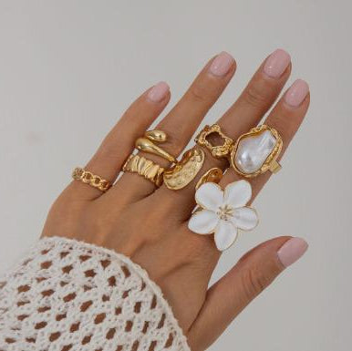 Hand wearing multiple gold rings with a white lace sleeve on a neutral background