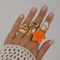 Hand wearing multiple gold rings with a focus on an orange floral ring against a neutral background