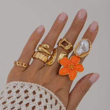 Hand wearing multiple gold rings with a focus on an orange floral ring against a neutral background